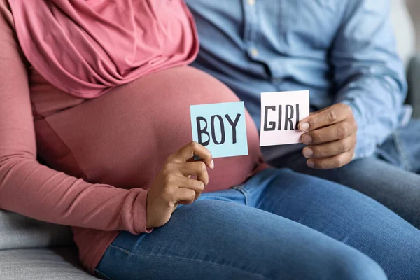 Pregnant couple holding boy and girl cards in pink and blue colors, unrecognizable future parents sitting on couch at home together, guessing gender of their upcoming baby, cropped, closeup shot