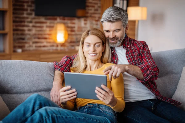 Smiling mature caucasian man hug lady and pointing finger at tablet, watch video in living room interior. New app, online shopping at home, chat and call at home, great offer and ad during covid-19