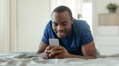 African American Guy Texting On Smartphone And Browsing Internet Lying On Bed Spending Weekend Morning In Modern Bedroom At Home. Gadgets And Mobile Communication Concept