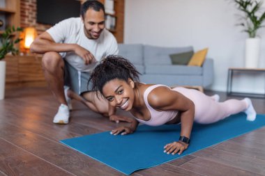 Cheerful millennial african american man in sportswear helping to wife doing push ups in living room interior. Love and relationship, fitness lesson, sports at home together and body care due covid-19