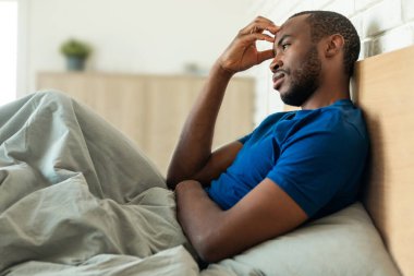 African American Man Thinking Touching Head Having Problems Sitting In Bed In Modern Bedroom At Home. Male Mental Health And Depression Concept. Side View