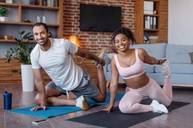 Smiling millennial african american man and woman in sportswear do stretching for legs on mat with bottle of water in room interior. Workout for muscle, fitness, sports at home together and body care
