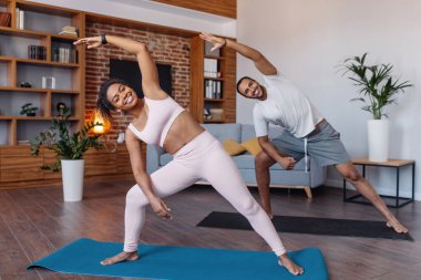 Happy millennial african american male and woman in sportswear doing body stretching in living room interior. Fitness, sports at home, workout together and health care during covid-19 quarantine