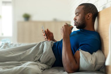 Black Man Using Smartphone Drinking Coffee Lying In Bed In Modern Bedroom At Home In The Morning. Millennial Male Texting And Browsing Internet Via Cellphone. Side View Shot
