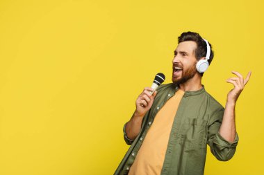 Joyful middle aged man in headphones listening to music and singing song into microphone, male in headset enjoying karaoke over yellow studio background, copy space
