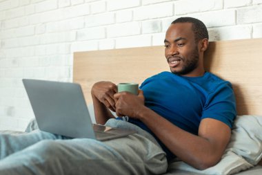 African American Man Using Laptop Watching Movie Online Drinking Coffee Lying In Bed In Modern Bedroom At Home. Guy Working On Computer In The Morning. Technology Lifestyle