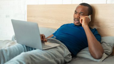 Discontented Black Freelancer Guy Talking On Cellphone Using Laptop Computer Working Online Lying In Bed At Home. Man Communicating Via Smartphone In Modern Bedroom. Distance Job Problems