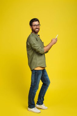 Positive caucasian man using smartphone and smiling at camera on yellow studio background, full length shot. Male with modern gadget chatting online, browsing web, posting in social media