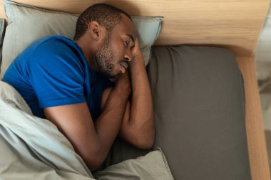 High Angle Shot Of Sleeping Black Guy Napping Holding Hands Near Face In Bed At Home. Male Resting In Modern Bedroom Enjoying Healthy Sleep. View From Above. Recreation Concept
