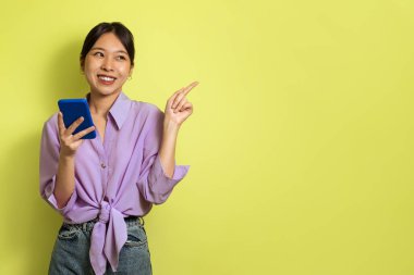 Happy Korean Woman Using Mobile Phone Pointing Finger Aside Advertising Mobile Offer Standing Over Yellow Background, Studio Shot. Technology And Gadgets. Empty Space