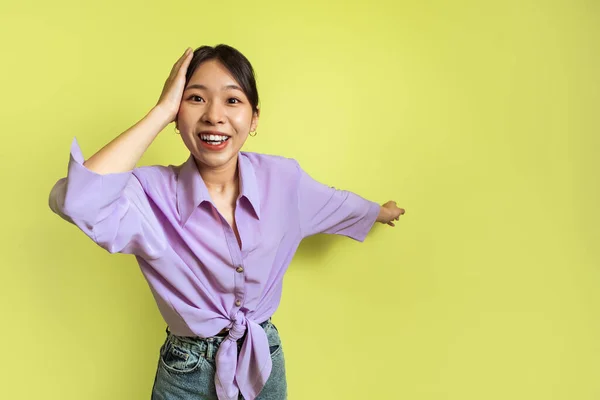 Excited Asian Female Pointing Aside Touching Head Advertising Wow Offer On Yellow Background In Studio. Woman Showing Copy Space Smiling To Camera. Look There Concept. Selective Focus