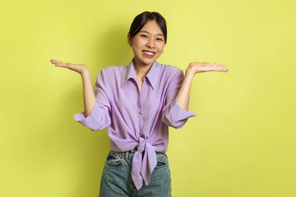 Asian Lady Shrugging Shoulders Smiling To Camera Standing Posing Over Yellow Background In Studio. I Dont Know Which Option Is Better, Comparison Concept