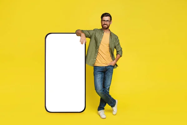 Full body length of man leaning on big smartphone with empty white screen, standing on yellow studio background, mockup. Mobile app advertisement