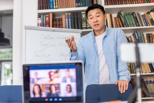 Online Teaching. Chinese Teacher Man Having Distance Class Making Group Video Call With Students Talking To Laptop And Phone Standing In Modern Classroom Indoor. Selective Focus On Tutor