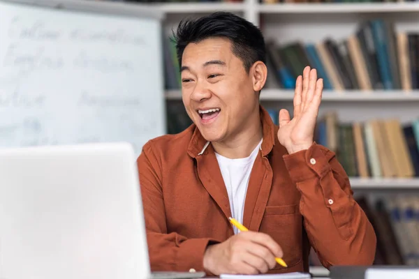 E-Teaching. Joyful Korean Male Teacher Making Video Call Waving Hello To Computer Teaching Remotely Sitting At Workplace Indoors. Modern Distance Education Concept. Selective Focus