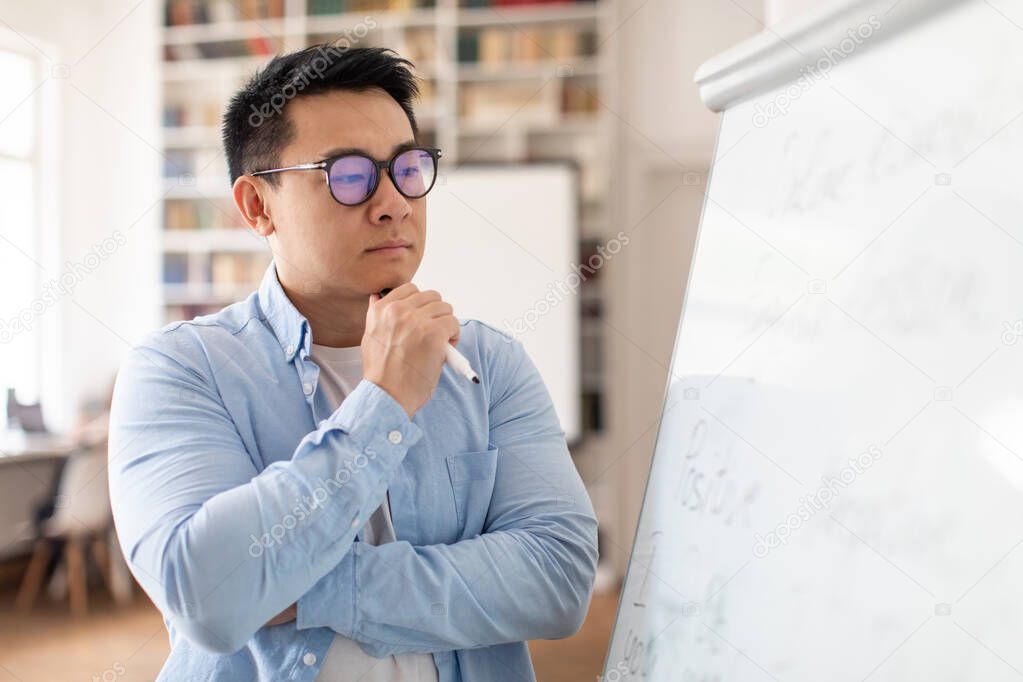 Thoughtful Asian Middle Aged Man Teacher Thinking Looking At Whiteboard Having Class Standing In Modern Classroom Indoors, Wearing Eyeglasses. Education And Science Concept. Selective Focus