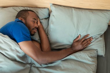 African American Millennial Man Sleeping Alone Touching Pillow Lying In Bed In Moodern Bedroom At Home. Healthy Rest And Sleep Concept, View From Above