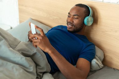 Afrifcan American Guy Wearing Headphones Using Smartphone Watching Mobile Video Lying In Bed In Modern Bedroom At Home. Male Using Application On Cellphone. Gadgets And Technology