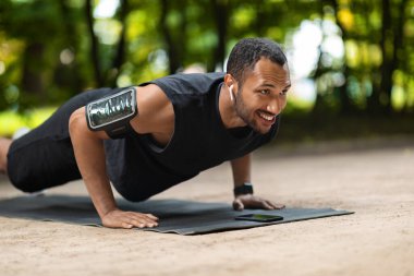Motivated handsome young african american guy sportsman planking at public park, exercising on fitness mat, looking at copy space and smiling, using earbuds and smartphone armband