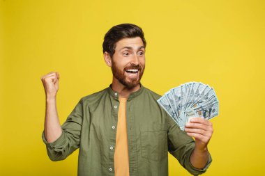 Overjoyed middle aged man holding lots of money, gesturing YES, male with fan of dollars showing casino or lottery win, achieving financial success over yellow studio background
