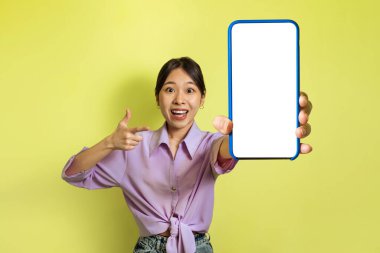 Excited Japanese Female Showing Large Smartphone With Blank Screen Pointing Finger Posing On Yellow Studio Background. Check This Mobile Offer Concept. Mockup