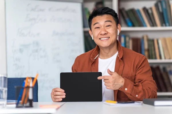 Cheerful Japanese Male Teacher Showing Digital Tablet Blank Screen Wearing Earbuds, Smiling To Camera Sitting At Workplace Indoor. Tutor Recommending E-Learning Website. Mockup