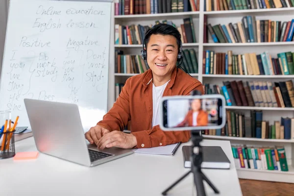 Online Education. Asian Male Tutor Making Video Lecture Via Cellphone And Laptop Wearing Headset Sitting At Workplace. E-Teacher Video Calling Having Distance Class Indoor. E-Teaching Concept