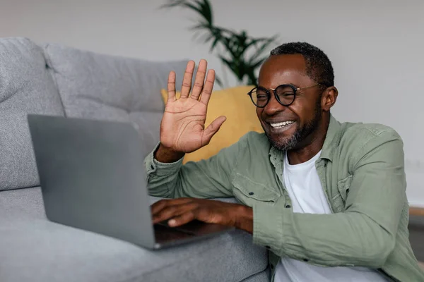 Smiling adult african american man in glasses waving hand gesture at laptop in living room interior. Work remote, business at home with modern technology, freelance and video call meeting due covid-19