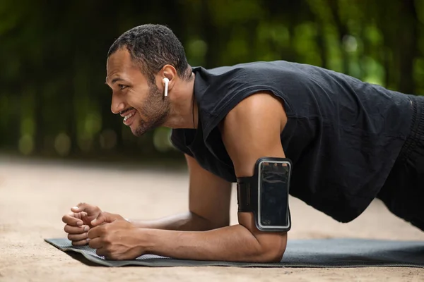 Side view of positive young black guy having workout outdoors, happy african american athletic man planking on fitness mat at public park, using earbuds listening to music, closeup