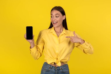 Great Mobile Application. Lady Showing Phone With Empty Screen Recommending New App Pointing Finger Standing In Studio Over Yellow Background. Technology And Gadgets