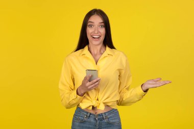 Happy Young Woman Using Mobile Phone Smiling To Camera Advertising Application Standing On Yellow Studio Background. Great App For Your Smartphone. Gadgets And Technology Concept