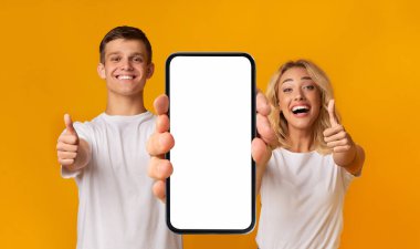 Cheerful excited young caucasian couple with open mouth show smartphone with blank screen and thumbs up, isolated on yellow background, panorama, studio. Great sale, new app, online ad and offer