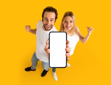 Happy millennial caucasian couple in white t-shirts enjoy winning online, show smartphone with blank screen, isolated on yellow background. Huge sale, ad and offer, website, presentation of new app