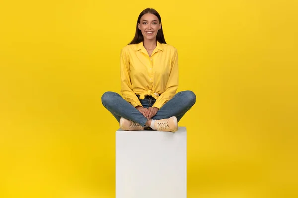 Happy Young Female Sitting On Cube Posing On Yellow Background In Studio, Smiling To Camera. Millennial Lady Expressing Positive Emotions Wearing Casual Clothes. Youth Fashion Concept