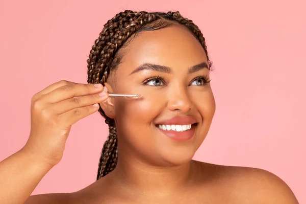 Portrait Of Happy African American Lady Applying Facial Serum Moisturizing Skin Posing Looking Aside Over Pink Background In Studio. Beauty And Cosmetics Concept