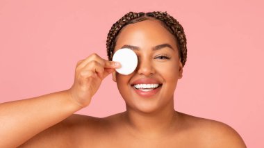 Facial Skincare. Pretty Overweight African American Lady Covering Eye With Cotton Pad Posing Smiling To Camera Over Pink Studio Background. Beauty And Skin Care Cosmetics Concept. Panorama