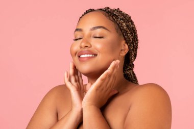 Facial Skincare. Portrait Of Happy Oversized Black Female Touching Face Caring For Perfect Smooth Skin Posing With Eyes Closed Over Pink Studio Background. Beauty And Selfcare Concept