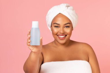 Happy Plump African American Lady Showing Cosmetic Bottle Recommending Great Body Lotion Posing Wrapped In Towel Over Pink Studio Background. Beauty Products Concept