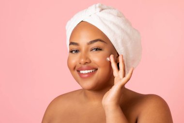 Headshot Of Obese African American Female Applying Moisturizer Cream Moisturizing Face Skin Posing Smiling To Camera Over Pink Studio Background. Facial Skincare Cosmetics Concept
