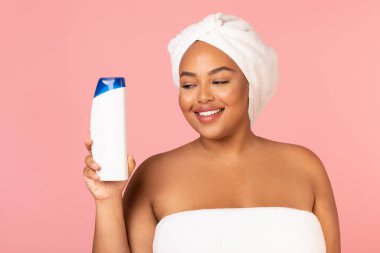 Cheerful Overweight Black Female Holding Shampoo Bottle Caring For Hair Posing With Wrapped Towel On Head Standing In Studio Over Pink Background. Hair And Body Care Cosmetics Concept