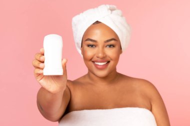 Happy Overweight Black Lady Showing Antiperspirant Stick Advertising Product For Sweat Protection, Standing Wrapped In Towel Smiling To Camera Over Pink Background. Studio Shot