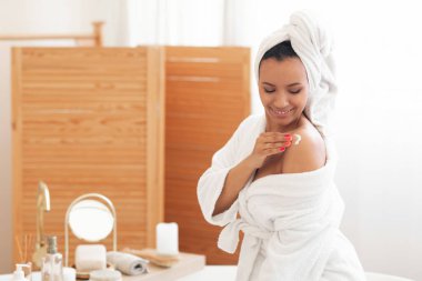 Bodycare Cosmetics. Pretty Mixed Lady Applying Body Lotion On Shoulders Moisturizing Skin Sitting In Modern Bathroom At Home, Wearing Bathrobe. Spa And Pampering Concept