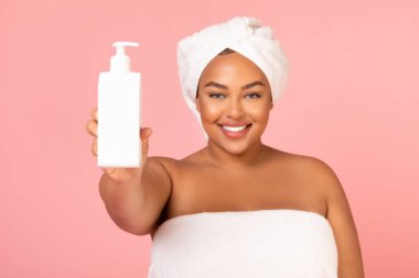 Overweight Black Woman Showing Dispenser Bottle Advertising Cosmetic Product Smiling To Camera Standing Wrapped In Towel Over Pink Background In Studio. Beauty Care And Spa Concept