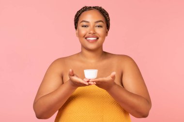 Plus Size African American Lady Showing Moisturizer Cream Jar Advertising Skin Care Cosmetics Smiling To Camera Standing Wrapped In Towel Over Pink Studio Background. Facial Skincare Concept
