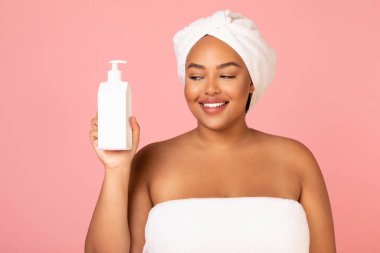 Happy Oversized Black Woman Holding Cosmetic Dispenser Bottle With Moisturizer Balm Standing Wrapped In Towel Over Pink Background In Studio. Skin And Body Care Concept