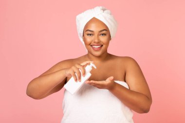Attractive Overweight African American Woman Applying Body Lotion Holding Cosmetic Bottle Smiling To Camera Posing Wrapped In Towel Over Pink Background, Studio Shot. Beauty And Pampering