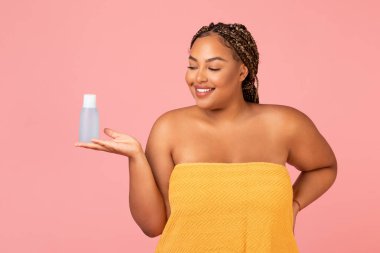Skincare Cosmetics. Oversized African American Female Holding Micellar Water Bottle Advertising Cosmetic Product Standing Posing Wrapped In Towel Over Pink Background. Studio Shot