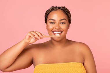 Obese Black Woman Brushing Teeth With Toothbrush Posing Standing Wrapped In Towel Smiling To Camera In Studio On Pink Background. Oral Hygiene And Healthy Teeth Concept