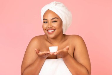 Happy Plump Black Female Holding Moisturizer Jar Recommending Skincare Cosmetics Smiling To Camera Posing Wrapped In Towel Over Pink Background. Studio Shot. Facial Skin Care Concept
