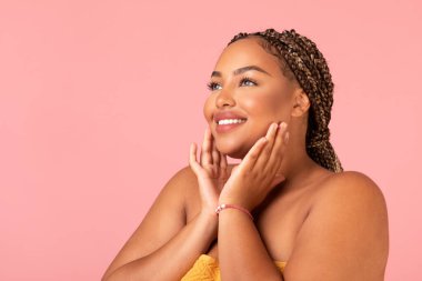 Plus Size African American Lady Touching Face Caring For Skin Smiling Looking Aside Posing Over Pink Background. Skincare And Beauty Routine Concept. Studio Shot